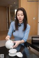 A woman in a blue dress pouring tea into a white teapot.