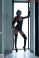 A woman in a black bodysuit and thigh high boots posing by a window.